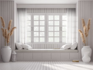 Large window with vintage style window seat in a room with white wood plank wall and floor and decorated with big white jar of dry reed flower.